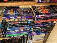 Photo showing a variety of Star Trek soft cover books stacked on shelves, titles visible include Star Trek Corona, Vulcan's Forge, The Lost Years, The Return, Enterprise The First Adventure, Engines of Destiny, and William Shatner's Log.