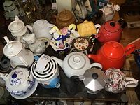 Photo showing multiple teapots in various shapes, colors and materials arranged on a table including a horse, red and orange pots, floral patterns and a moka pot