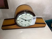 Front view of vintage mantle clock showing the wooden case, clock face with glass cover, and dial with Arabic numerals and logo.