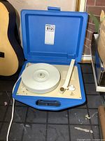 Record player inside open blue portable case showing white platter, tonearm, and control knobs.