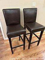 Photo showing two brown leather bar stools with black painted wooden legs and frames, cushioned seats and backs.