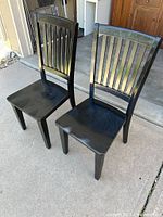 Two black painted wooden dining chairs photographed outside, showing vertical slatted backs, flat seats, and straight legs with some surface wear.