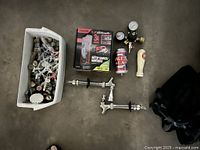 Photo shows a CO2 gas regulator with two pressure gauges and brass connectors, two stainless steel beer faucets with black shanks, two beer tap handles (white ceramic and cream bottle cap shape), a boxed CO2 cartridge charger labeled '3A', a beer can, and a large container filled with assorted bottle openers and keys.