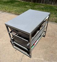 Photo of a gray metal frame office organizer with three black wire mesh drawers and a light gray wooden top placed outdoors on a concrete surface.