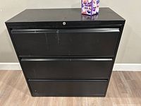 Front view of black 3-drawer lateral file cabinet with lockable top drawer, showing scratches and wear on surface.