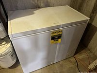 Frigidaire white chest freezer showing top lid, front EnergyGuide label and manual temperature control dial knob on right bottom corner, placed on concrete floor.