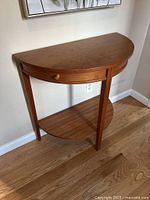 Photo of a half-round wooden side table with a drawer and open shelf placed against a wall on hardwood flooring.