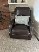 Dark brown leather recliner chair with a gray textured pillow placed in the seat. The chair is positioned on a carpeted floor against a wall with a brick fireplace partially visible to the left.