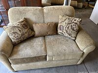 Beige upholstered love seat with two large floral pattern throw pillows and one smaller rectangular pillow, carpeted warehouse floor background.