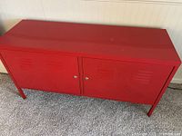Red metal cabinet with double doors, placed on carpet, suitable for foot or side of bed storage.