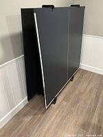 Photo showing folding black Olhausen ping pong tabletop stored vertically against a wall, highlighting the fold and wheels for rolling storage.