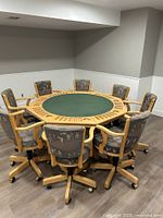 Photo showing an octagonal wooden poker or game table with green felt surface in the center and 8 rolling chairs around it in a room with wooden floor and two-tone gray-white walls.