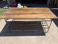 Photo of a rectangular coffee table placed outdoors on pavement. The table has a natural wood plank top and a black metal frame base with horizontal bars providing structure.