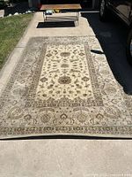 Photo showing a large rectangular beige and brown area rug with floral and geometric patterns placed on a concrete driveway with a small table and grass in the background.