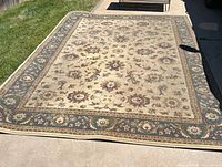 Full view of the large rectangular area rug laid out on a concrete surface, showing the floral medallion pattern and blue border