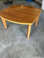 Square wooden coffee table with rounded edges, medium wood tone top and lighter legs, photographed on concrete floor.