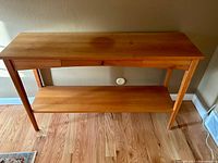 Front-facing photo of the natural wood sofa table showing its rectangular top, single drawer with knob, and lower shelf, placed on light wood flooring with a wall outlet behind.