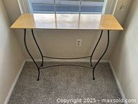 Photo showing rectangular light wood sofa table with black metal curved legs placed in front of a window on a carpeted floor.