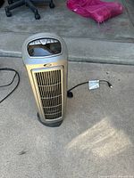 Full view of silver-gray vertical tower space heater with vent grille and digital control panel on top, power cord attached and unplugged, placed on concrete floor.