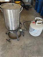 Turkey fryer kit consisting of large stainless steel pot with lid on metal burner stand connected to a propane tank with hose and control valve.