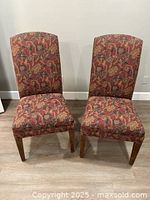 Two high back chairs with patterned red and orange fabric upholstery, wooden legs, positioned on a hardwood floor against a white wall.
