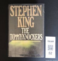 Front cover of The Tommyknockers novel by Stephen King showing the black dust jacket with beige title lettering and glowing effect feature at bottom.