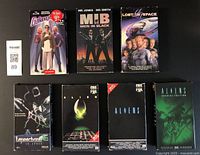 Front view of 7 science fiction VHS tapes arranged in two rows, showing titles and artwork including Galaxy Quest, Men in Black, Lost in Space, Leprechaun 4 in Space, Alien, and Aliens.