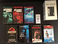 Photo showing nine VHS tapes and a Die Hard Trilogy box set arranged on a surface. Titles visible include Beneath the Planet of the Apes, Planet of the Apes, The Poseidon Adventure, Jurassic Park, Total Recall, Bloodsport, Cliffhanger, Die Hard Trilogy box set.