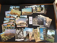 Photo showing a spread of 61 assorted vintage postcards laid out on a dark surface. Various images including landscapes, architecture, and city scenes. Visible mix of color and black and white postcards.