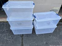 Five clear medium size plastic storage containers stacked outside against a wall, three on left stacked, two on right stacked. Some containers marked with numbers '48' and '49'. All containers have lids, two with blue latch handles.