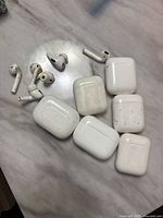 Multiple AirPods 1st generation and AirPods Pro earbuds scattered with six charging cases on a marble surface, showing wear and dirt marks on charging cases.
