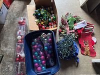 View of multiple containers including a blue plastic bin filled with various Christmas ornaments in purple, blue, and silver, a cardboard box with red and gold ornaments, two Christmas stockings with festive designs, and a coil of string lights.