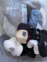 Photo showing a pile of white, black, and beige hats, along with blue denim pants and other clothing items inside a container.