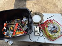 Plastic container filled with assorted tools including orange handled levels, hammer, pliers, and tape measure. White and yellow coiled cables and an electrical device are placed beside the container.