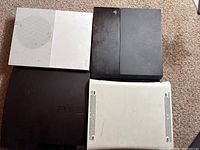 Top-down view of four game consoles: white Xbox 360, black PS4, black PS3, and white Xbox 360 on carpeted floor showing surface wear and discoloration.