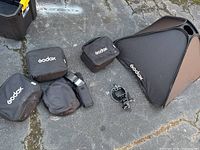 Overview of Godox camera equipment including large softbox, flash units in carrying cases, round pouch, and mounting bracket on pavement.