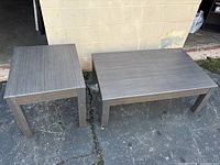 Two gray living room tables outdoors on pavement next to a beige wall, a larger coffee table and a smaller side table with rectangular tops and four legs each.