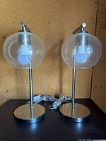 Pair of Howin glass globe table lamps side by side, showing front view of lamps with visible clear glass globes, metal stands and round bases.
