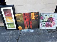 Three large framed wall art paintings placed on ground outside, showing floral and nature themes in red, orange, green, white, and pink colors.