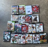 Photo showing 18 video game cases for Xbox 360, Wii, and PlayStation 2, including titles like Sims 2, NBA Live 07, NBA 2K series, Madden NFL series, Call of Duty, Dance Dance Revolution, and others.