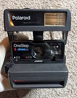 Front view of the black Polaroid 600 OneStep CloseUp camera showing the flash, lens, viewfinder, and branding.