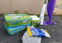 Purple Swiffer mop handle with flat mop head connected to several boxes of Swiffer Sweeper dry sweeping cloth refills and a pack of disposable wipes all displayed outdoors on pavement.