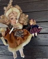 Photo showing both dolls, with the larger Tiffany Victorian doll in front wearing a brown and fur outfit and the smaller doll behind in a blue and red dress.