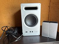 White Vizio subwoofer with front-facing driver, slot port, and control buttons. Cables visible behind subwoofer.