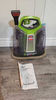 Front view of the green and black Bissell Little Green ProHeat Pet steam cleaner with manual in front on floor.
