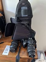 Nikon DSLR camera body with attached 18-55mm lens, additional lens and two camera bags displayed on wooden surface.