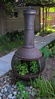 Full view of metal chiminea stand in garden setting showing tall chimney neck with embossed swirl patterns and mesh base with plants inside.