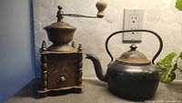 Front view of a vintage metal coffee grinder and heavy metal kettle on a countertop.