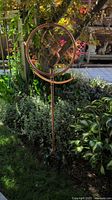 Full view of one copper decorative sprinkler stand inserted in lawn with plants in background showing circular frame and decorative spinning blades.