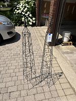 Two brown metal garden trellises on paved driveway, showing overall shape, size, and pointed finials.